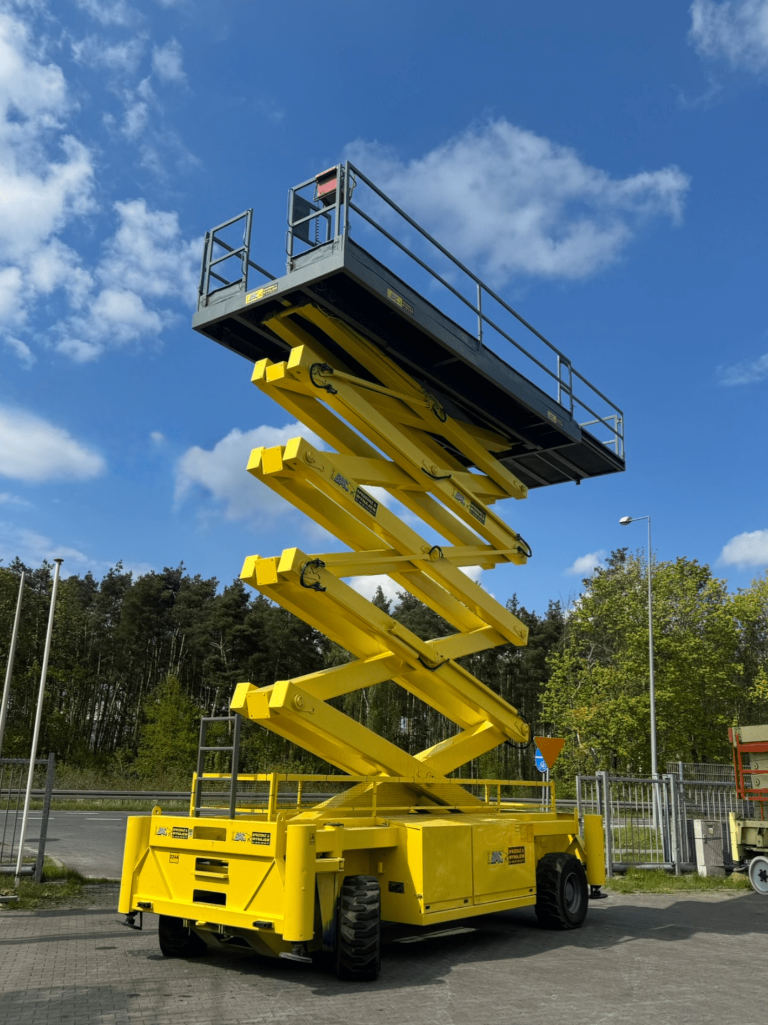 Podnośnik nożycowy Holland Lift Combistar M250DL27-4WD/p/n o wysokości roboczej 27 metrów, z wysuniętą platformą i napędem 4x4, gotowy do pracy na placu budowy