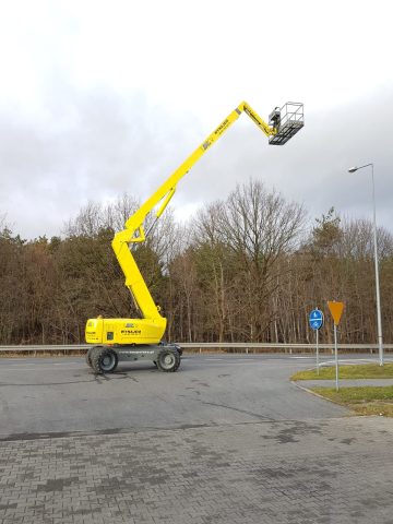 A basket lift in the parking lot next to the trees.