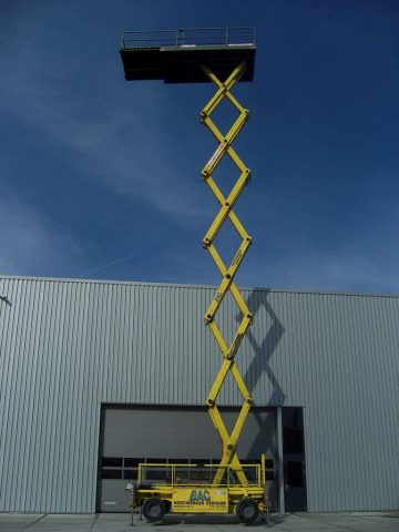 Желтый ножничный подъемник перед промышленным зданием.