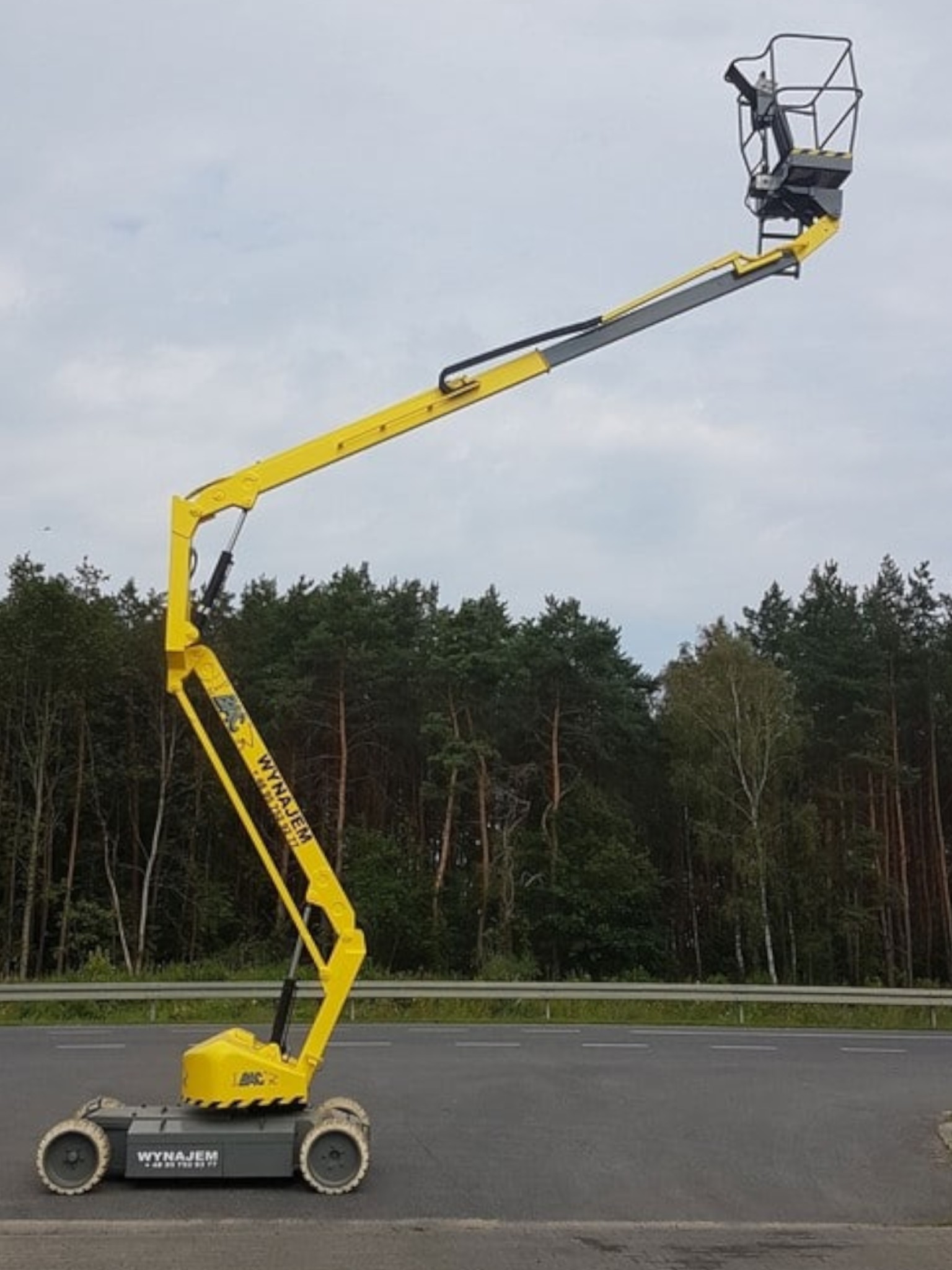 Żółty podnośnik koszowy obok drogi i lasu.
