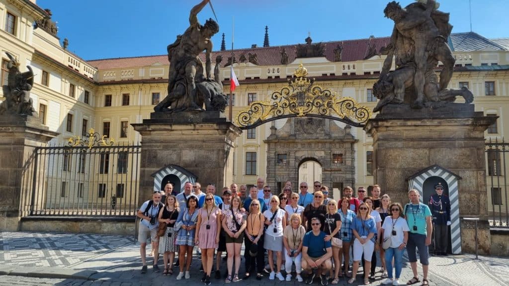 Skupina turistů před palácem s bránou a sochami.