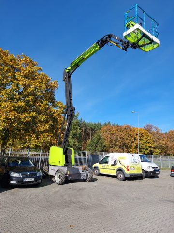 Podnośnik koszowy na parkingu, jesienne drzewa w tle.